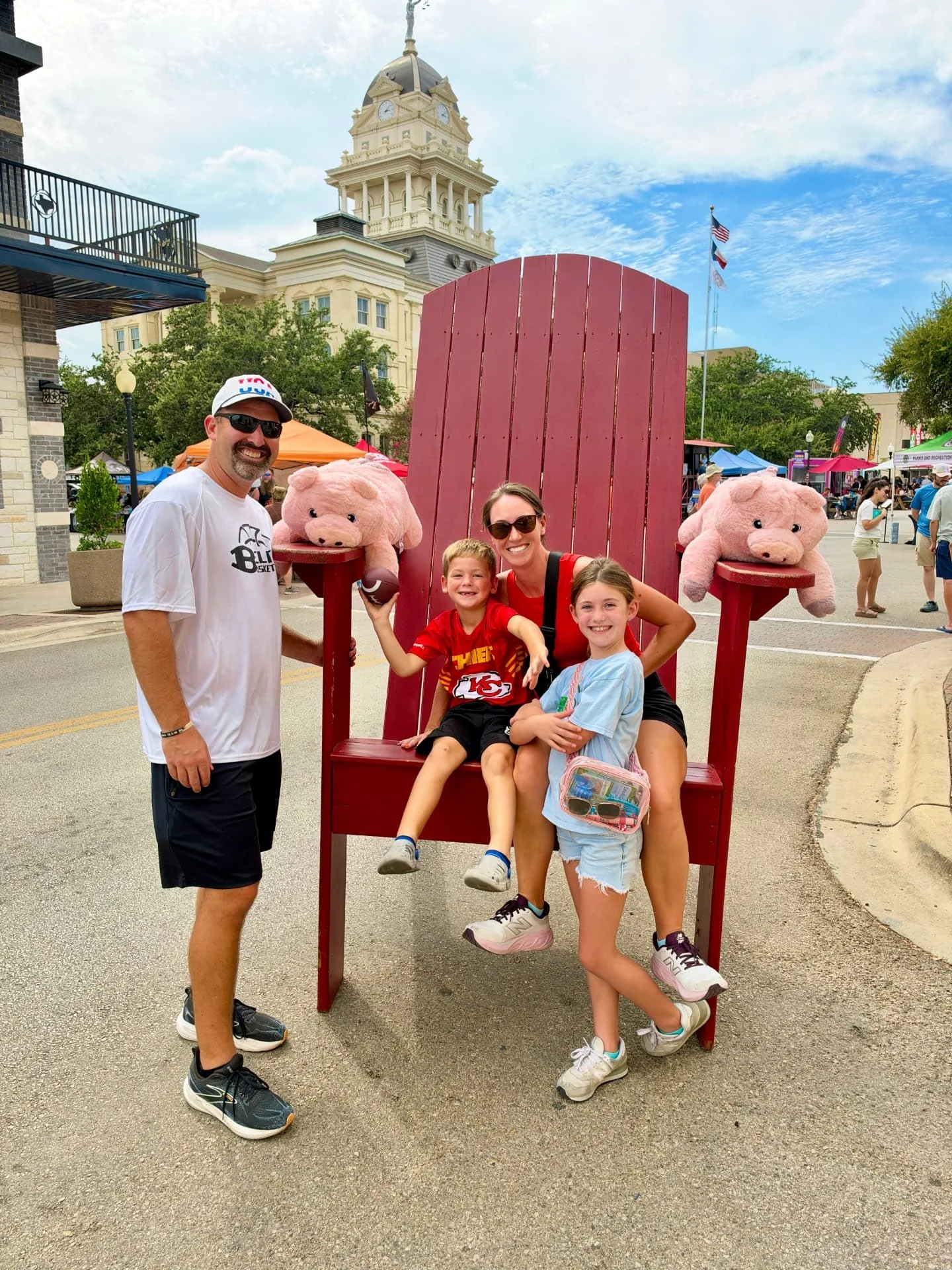A family poses in an oversized red chair on a downtown Belton street during a festival, with vendor tents and the courthouse tower behind them